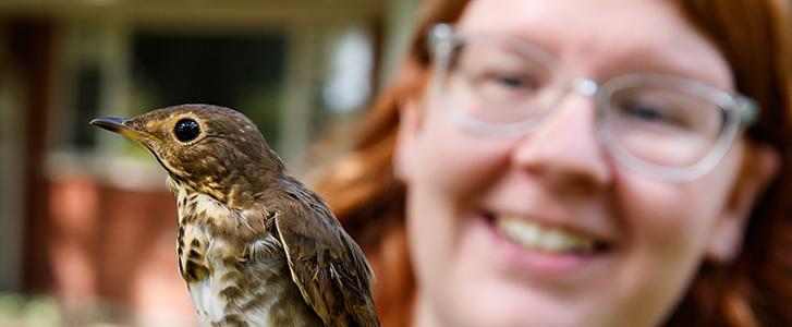 Small bird in focus, smiling woman with glasses blurred in background.