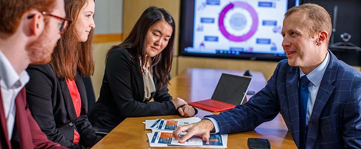 Person points to documents on a table while talking to a group of people, with a screen displaying graphs in the background..