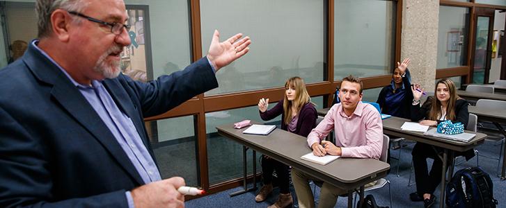 Professor talking to students in a classroom; three students raise their hands.