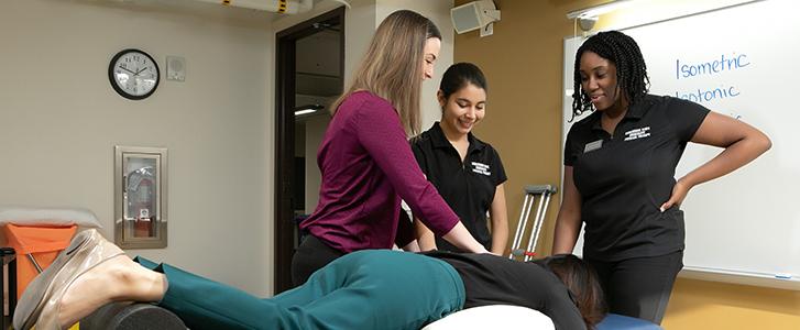 A physical therapy student performs manual therapy on a peer while two other physical therapy students observe in a classroom.