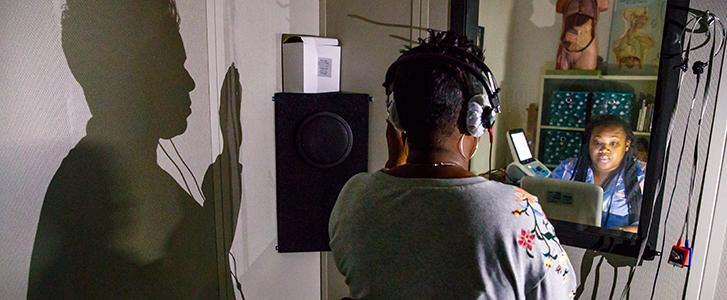 Person with headphones facing a window where another person is typing on a laptop on the other side.