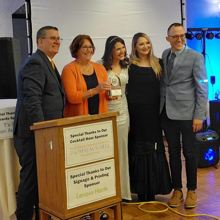Five smiling people, one holding an award, stand next to a podium at an event.