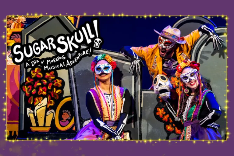 Three performers in colorful Día de Muertos costumes pose with a skull prop in front of tombstone scenery. The Sugar Skull! logo appears on the left.
