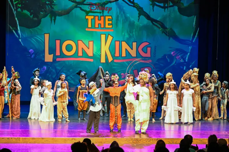 Cast of The Lion King musical on stage, performers in colorful costumes, large logo in background.