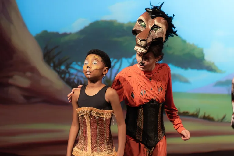 Young performer with face paint looks up at Scar-masked actor on stage.