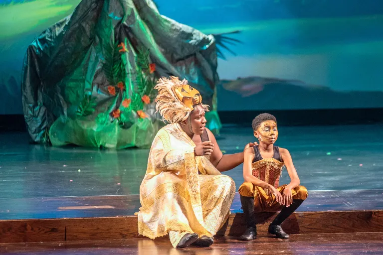 Teen in golden costume comforts a child in theatrical costume on stage.