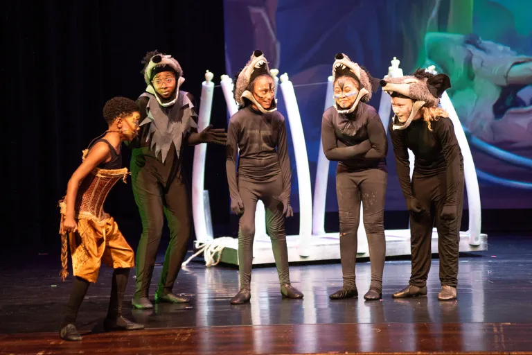 Five children performing on stage. One in gold, four in grey hyena costumes look at him intently.