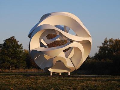 White, abstract, spherical sculpture with intertwining ribbons in a grassy field at sunset.