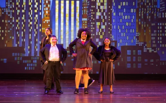 Four performers pose confidently at center stage with a city skyline backdrop behind them