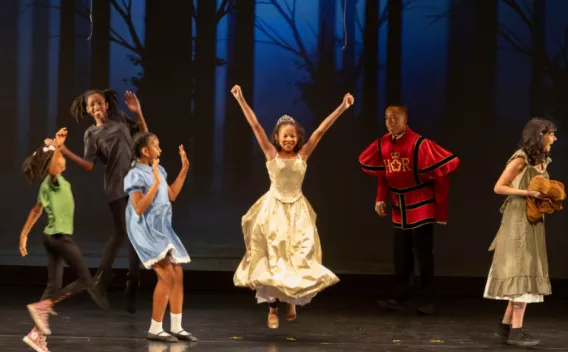  Performers dressed in various costumes dance onstage, with one performer in a gown leaping in the center